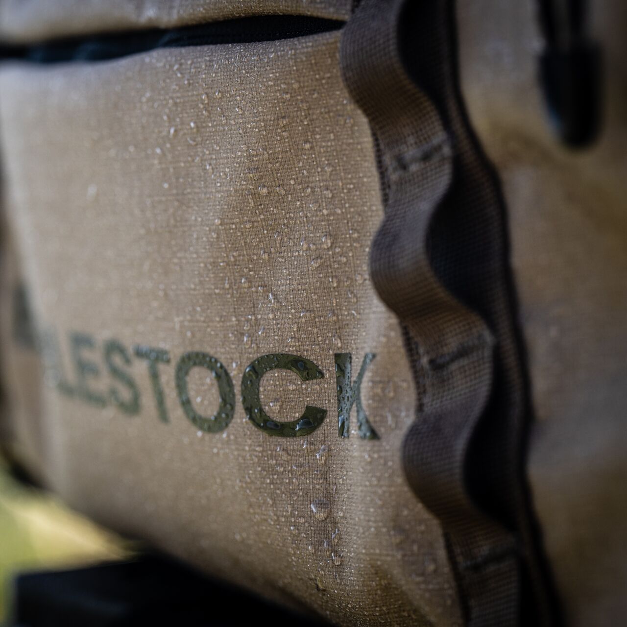 Nahaufnahme der EBERLESTOCK Hyllus Duffel 45, einer hellbraunen 45-Liter-Seesacktasche mit Wassertropfen auf ihrer robusten, strukturierten Oberfläche. Ein Teil des Wortes STOCK ist auf der Tasche zu sehen, die mit dunklen Besätzen für einen outdoor-tauglichen Look sorgt.