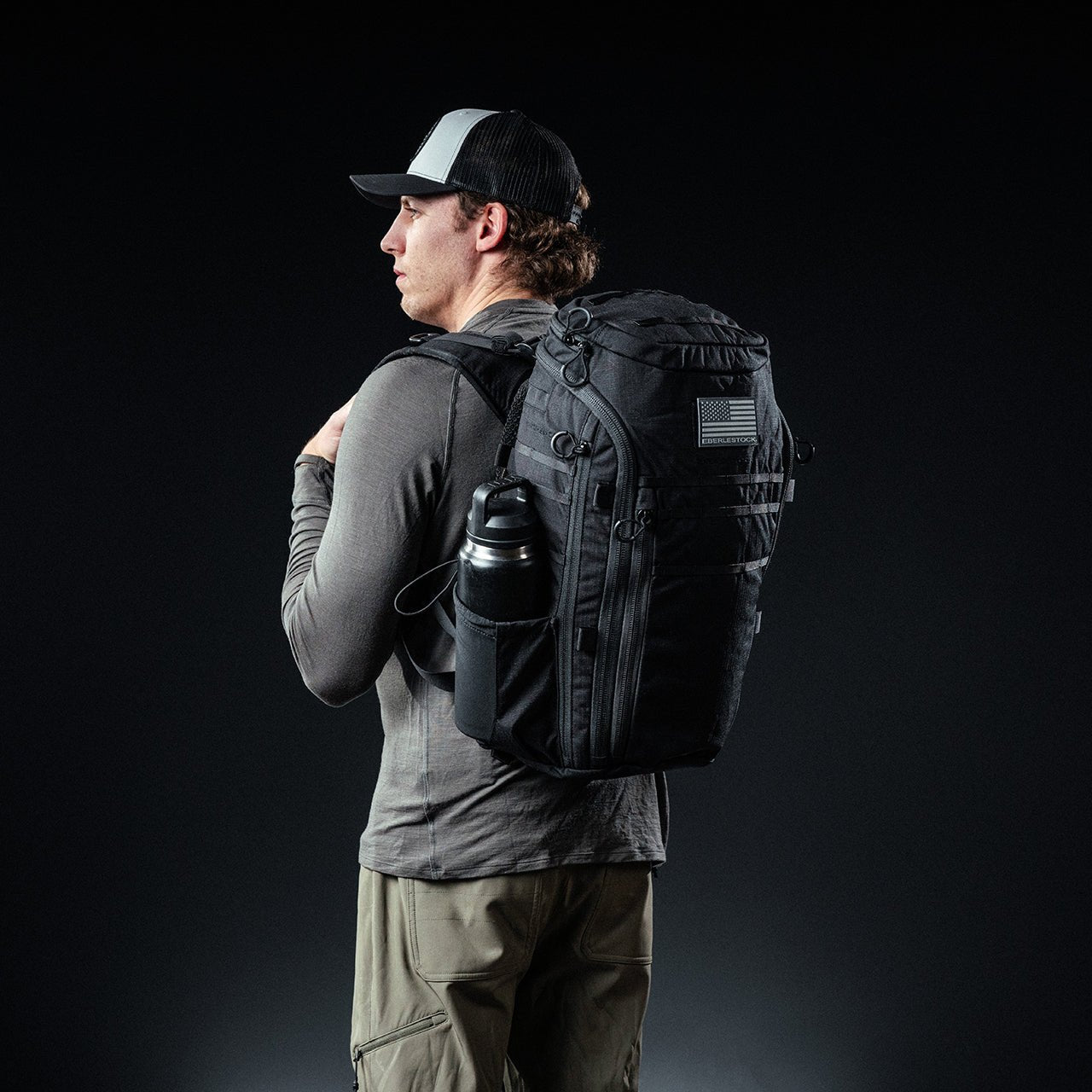 Eine Person in grauem Hemd, khakifarbener Hose und schwarz-weißer Mütze steht seitlich mit einem EBERLESTOCK Switchblade Pack - einem taktischen Rucksack aus 500D-Nylon - mit einer Wasserflasche und einem Aufnäher der amerikanischen Flagge vor einem dunklen Hintergrund.