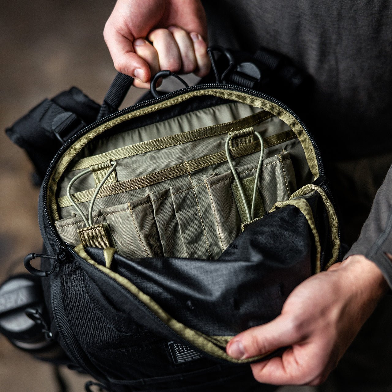 Eine Person hält einen EBERLESTOCK Switchblade Pack auf und gibt den Blick frei auf ein geordnetes Innenleben mit braunen Trennwänden, Taschen und elastischen Riemen. Der Rucksack ist aus strapazierfähigem 500D-Nylon gefertigt und eignet sich für den Transport von Ausrüstung und Equipment.