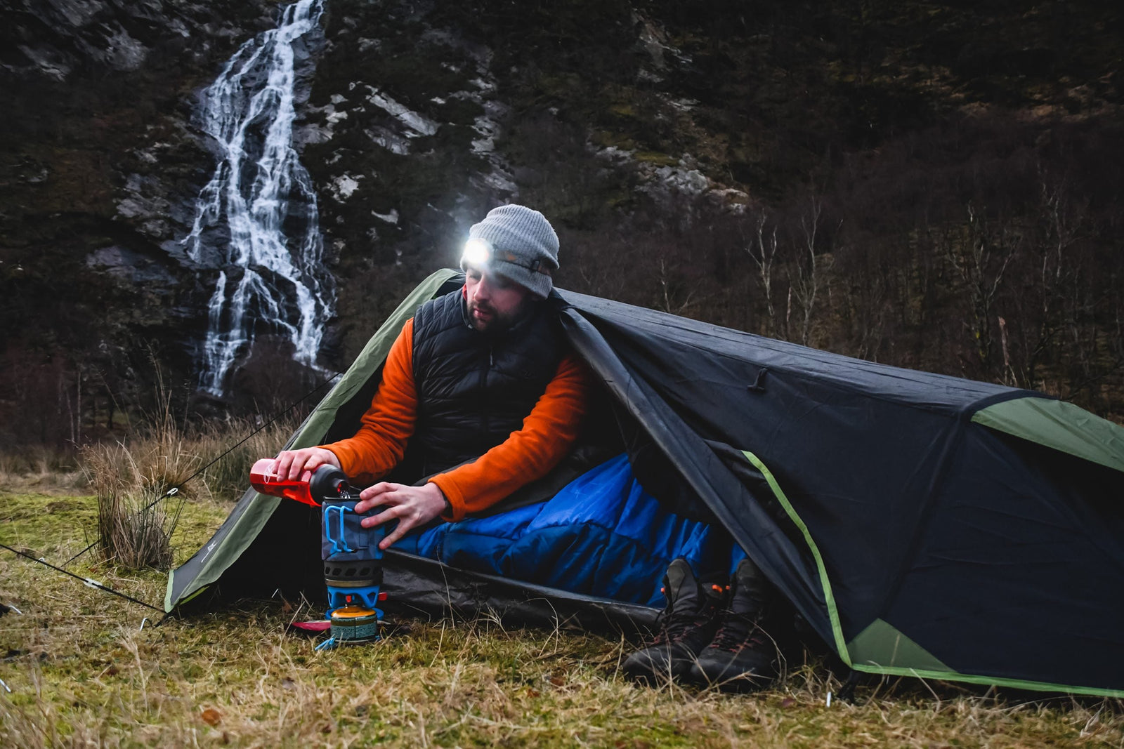 Mit Stirnlampe und Mütze sitzt eine Person in einem HIGHLANDER FORCES Highlander Zelt Blackthorn 1+ Gen2 - HMCT und benutzt einen Campingkocher neben sich, während im Hintergrund ein großer Wasserfall den felsigen Hang hinunterstürzt.