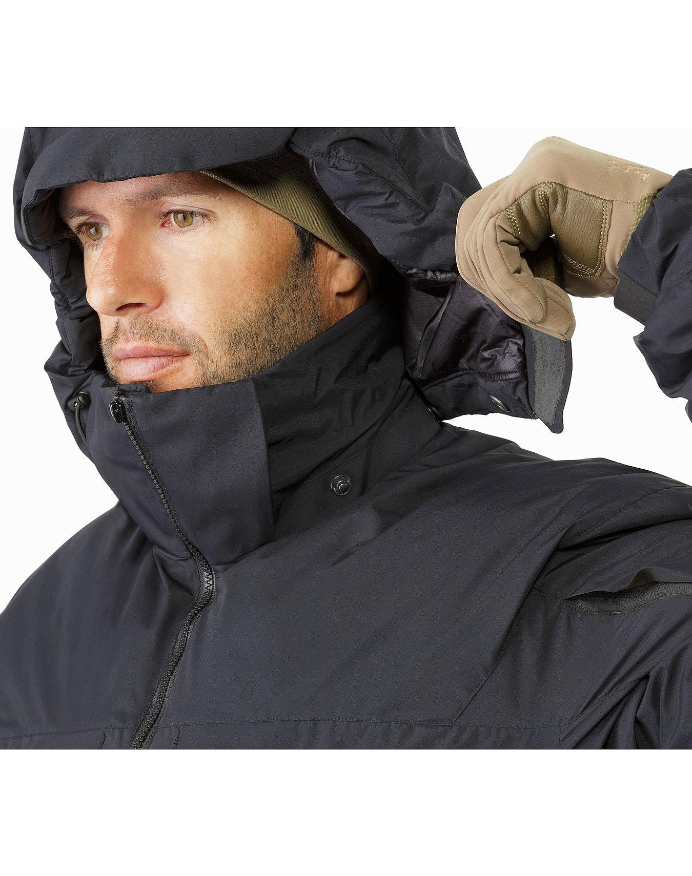 Ein Mann in einer schwarzen ARC'TERYX LEAF Cold WX Jacket SV Gen1.4 mit hellbraunen Handschuhen rückt seine Kapuze zurecht. Er trägt eine dunkle GORE WINDSTOPPER Innenkappe und schaut mit neutraler Miene zur Seite.