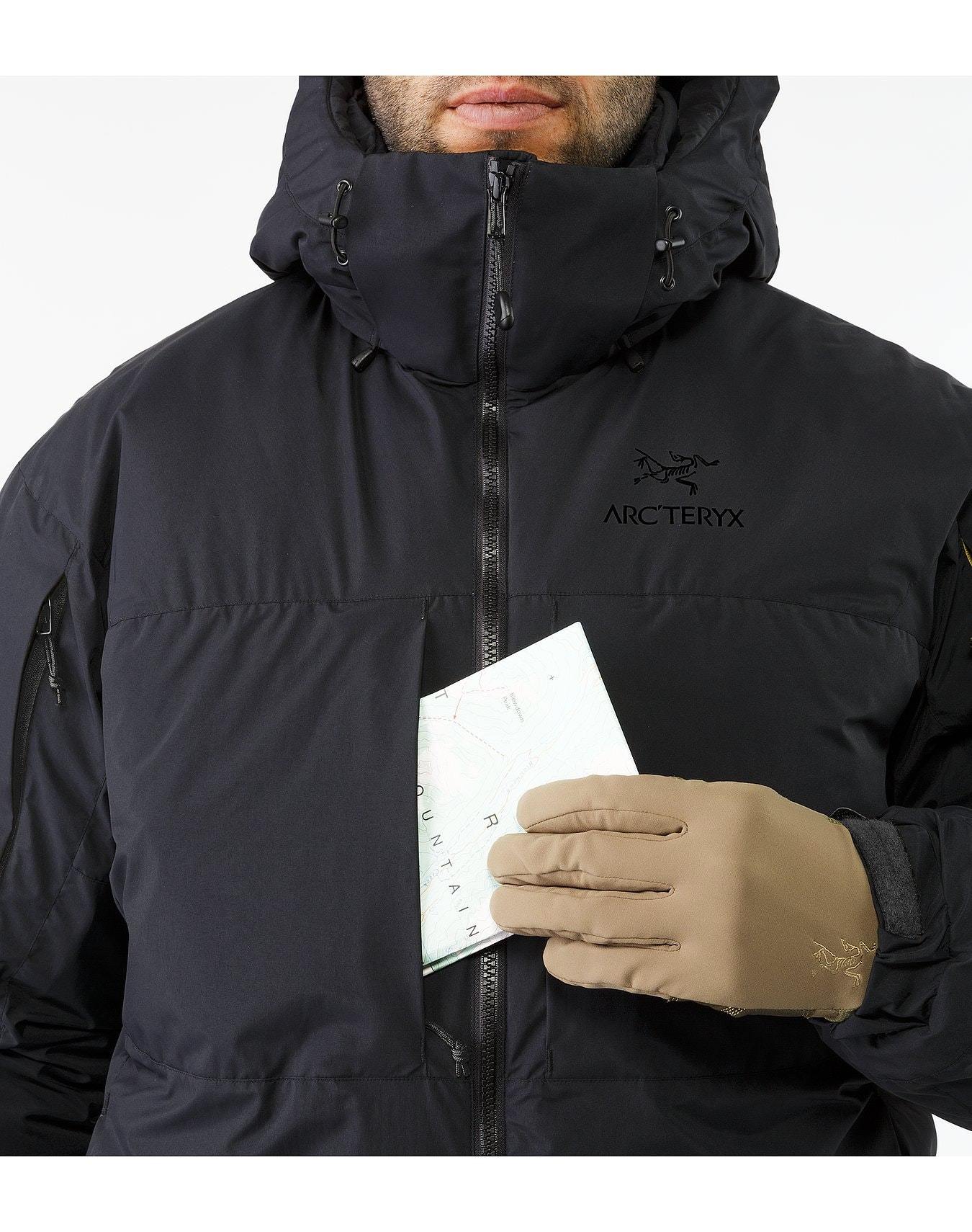 Eine Person in einer schwarzen ARC'TERYX LEAF Cold WX Jacket SV Gen1.4 mit hochgezogener Kapuze hält eine gefaltete Landkarte, die teilweise in der Brusttasche steckt; hellbraune Handschuhe bedecken die Hände und nur das untere Gesicht ist sichtbar.