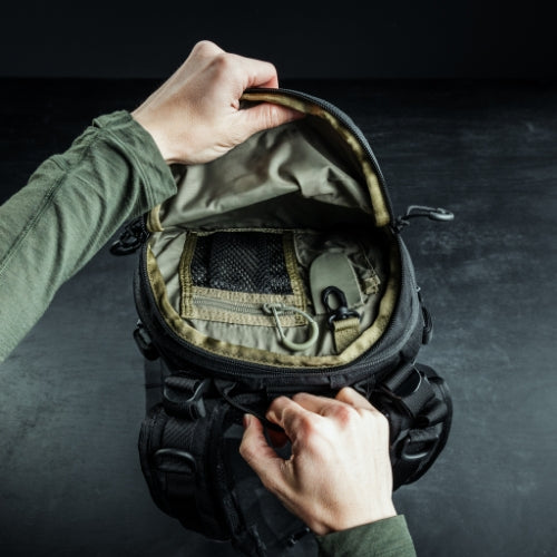 Eine Person in einem grünen langärmeligen Hemd öffnet einen EBERLESTOCK Bandit Pack und enthüllt sein organisiertes Inneres mit Netztaschen, Ausrüstung und einem MOLLE-Element vor einem dunklen Hintergrund.