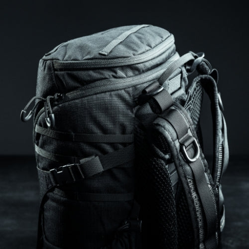 Eine Nahaufnahme des EBERLESTOCK Bandit Pack, eines schwarzen, robusten Rucksacks mit mehreren Reißverschlüssen, Riemen und MOLLE-Panel, vor einem dunklen Hintergrund. Das robuste Design macht ihn ideal für Outdoor-Abenteuer oder Reisen.