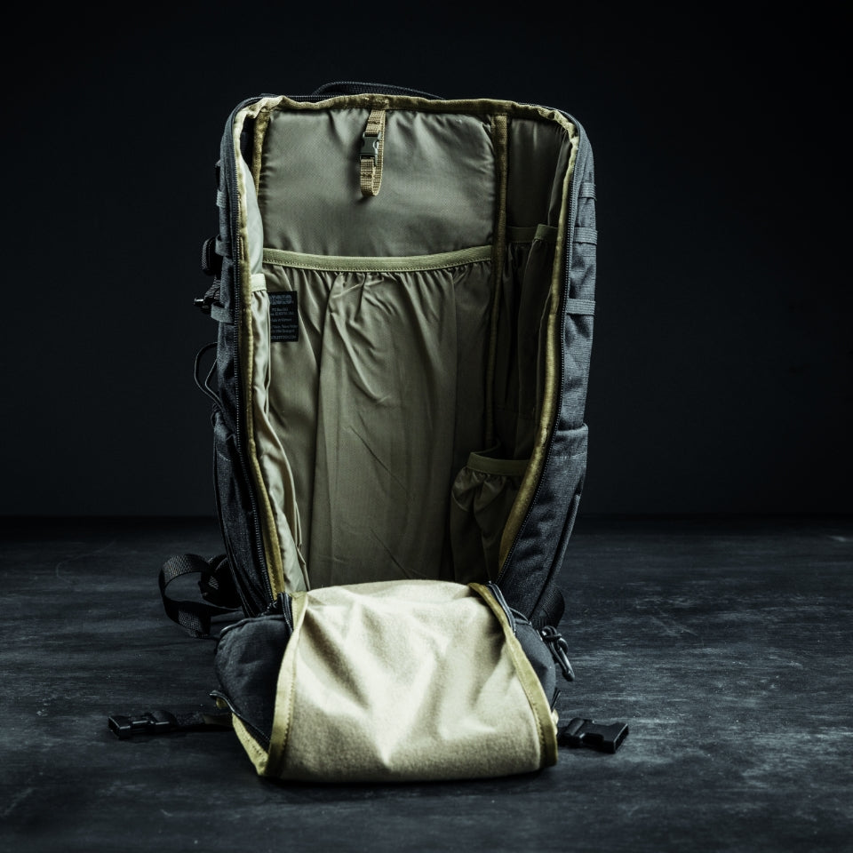 Ein EBERLESTOCK Bandit Pack steht aufrecht auf einer dunklen Fläche, der Reißverschluss ist geöffnet, um seinen geräumigen, leeren Innenraum mit zahlreichen Taschen und Fächern, einschließlich eines MOLLE-Panels, vor einem schattigen Hintergrund zu präsentieren.
