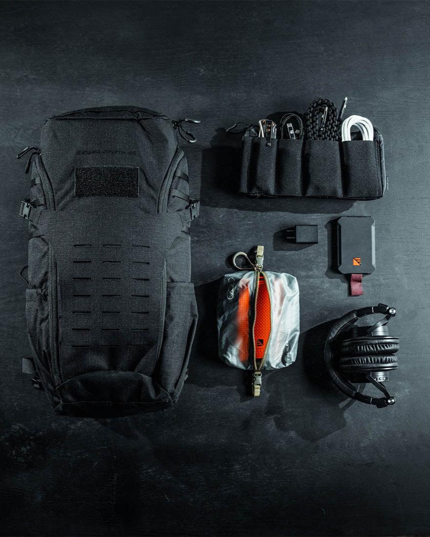 Eine flache Anordnung von schwarzer taktischer Ausrüstung auf einer dunklen Oberfläche, mit dem EBERLESTOCK Bandit Pack, einer technischen Organizertasche mit MOLLE-Paneel, Kabeln, einem tragbaren Ladegerät, einer kleinen Tasche und schwarzen Kopfhörern.