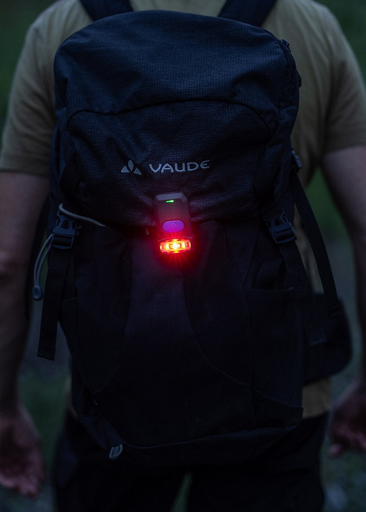 Eine Person mit einem schwarzen Vaude-Rucksack, an dem eine NORDRIDE Nordride Clipleuchte Scout befestigt ist, steht bei schwachem Licht im Freien. Der Fokus liegt auf dem Rucksack und der Außenbeleuchtung, wobei die Arme der Person teilweise sichtbar sind.