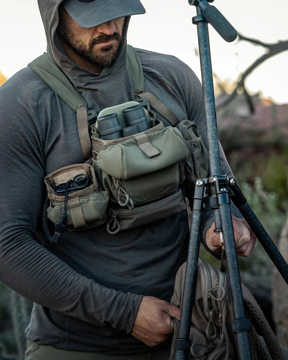 Eine Person mit grauem Kapuzenpulli und Mütze stellt im Freien ein Stativ ein, während sie ein EBERLESTOCK Bravo Modular Bino Pack Small mit Fernglastasche und Ausrüstungsfächern trägt.