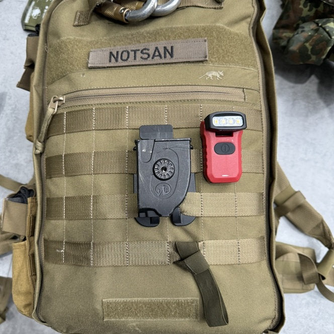 Ein hellbrauner taktischer NOTSAN-Rucksack mit einem schwarzen Clip und einer roten NORDRIDE Clipleuchte Paramedic Light auf der Vorderseite ruht auf einer hellen Fläche, wobei im Hintergrund teilweise eine militärische Tarnkleidung zu sehen ist.