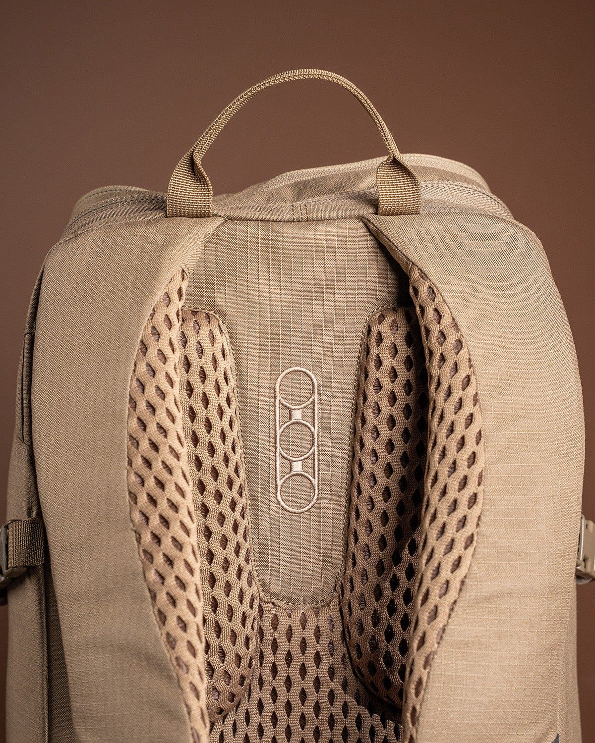 Eine Nahaufnahme des Rückenteils und des oberen Griffs des EBERLESTOCK Brute Scout Rucksacks zeigt gepolstertes, atmungsaktives Netzgewebe und drei ineinander greifende Ringe in der Mitte. Der Hintergrund ist einfarbig braun.