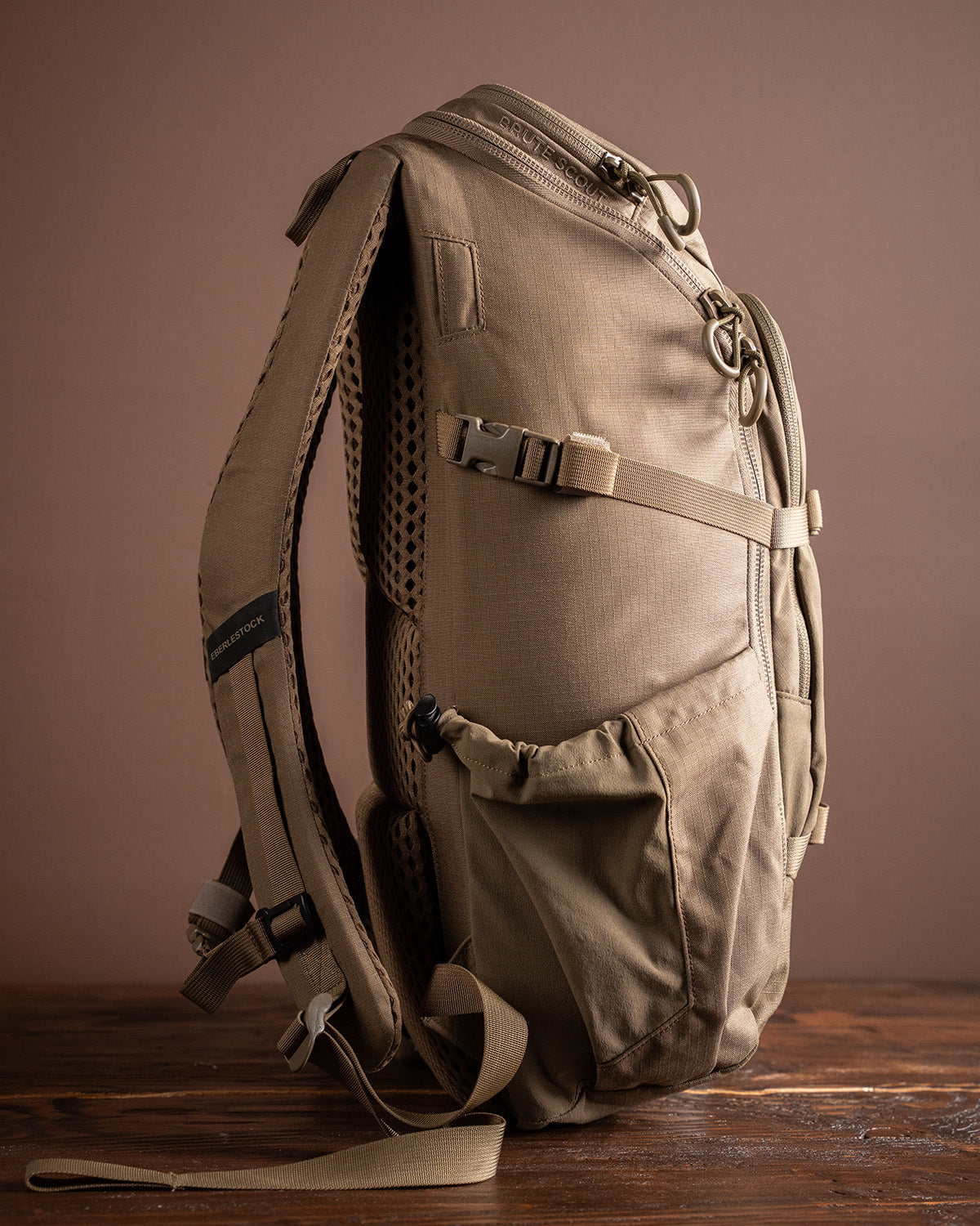 Der EBERLESTOCK Eberlestock Brute Scout ist ein hellbrauner taktischer Rucksack mit mehreren Reißverschlusstaschen, gepolstertem Netzrücken, verstellbaren Gurten und Schnallen. Er ist aufrecht auf einer hölzernen Oberfläche mit einem schlichten braunen Hintergrund abgebildet.
