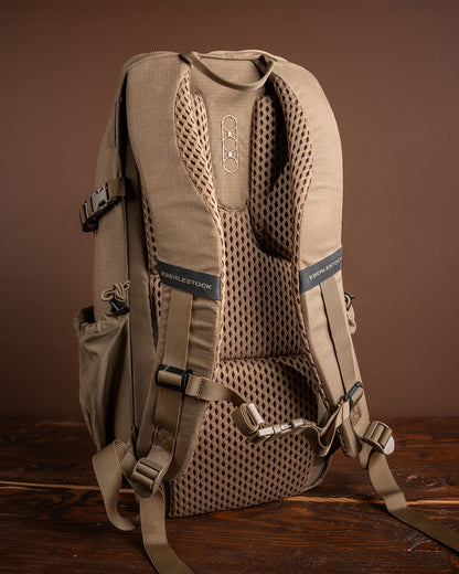 Der EBERLESTOCK Eberlestock Brute Scout in Hellbraun wird von hinten gezeigt und hebt die gepolsterten Mesh-Schultergurte, die verstellbaren Schnallen und die Seitentaschen vor einem braunen Hintergrund auf einer Holzoberfläche hervor.