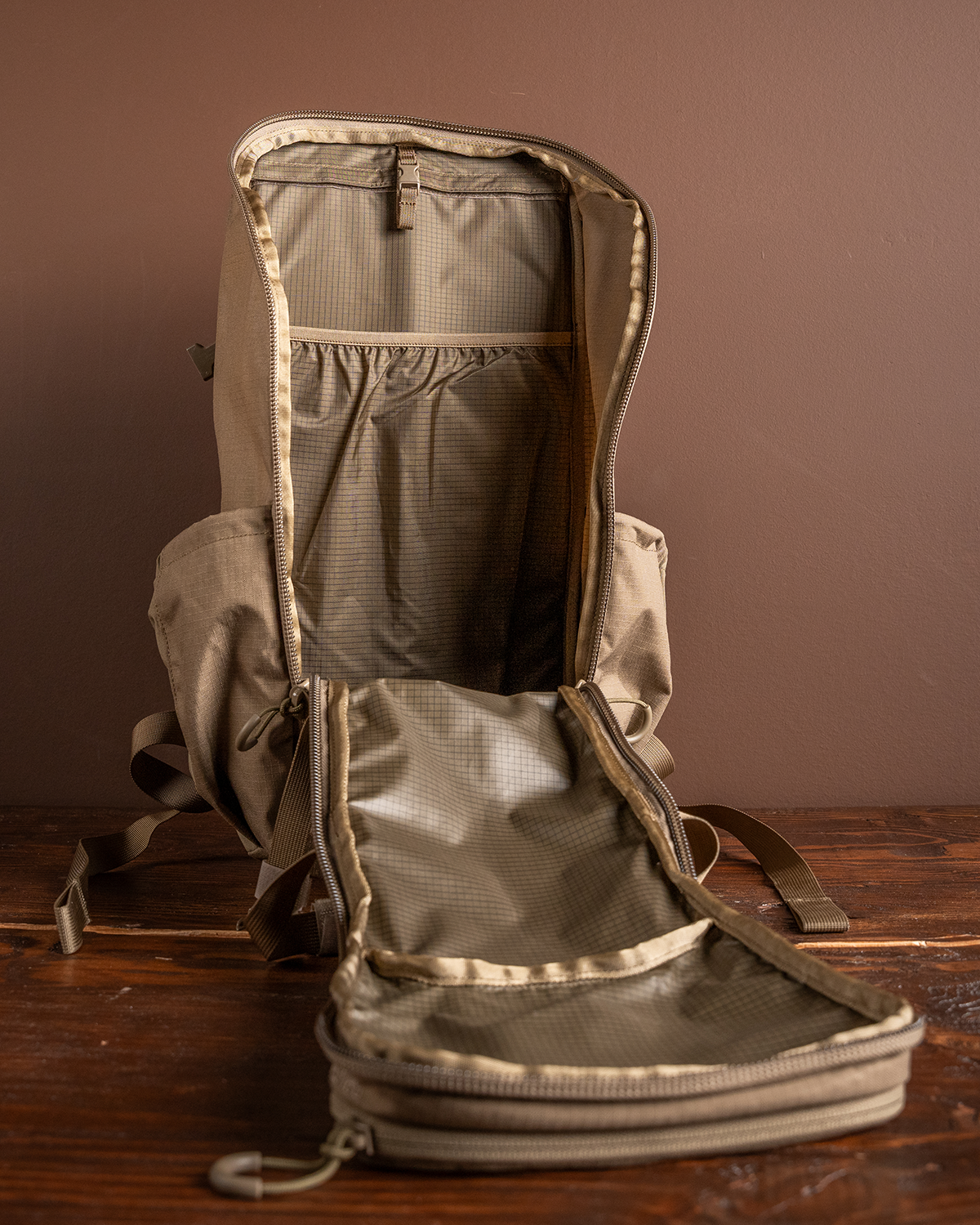 Ein hellbrauner EBERLESTOCK Brute Scout Rucksack steht aufrecht auf einer hölzernen Fläche an einer braunen Wand. Der Reißverschluss ist vollständig geöffnet, so dass das leere Hauptfach und die innere Netztasche zum Vorschein kommen.