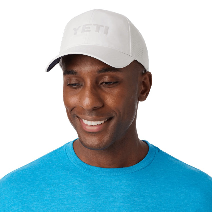 Ein lächelnder Mann trägt eine weiße YETI® Structured Performance Cap und ein hellblaues T-Shirt und blickt leicht nach unten vor einem schlichten weißen Hintergrund.
