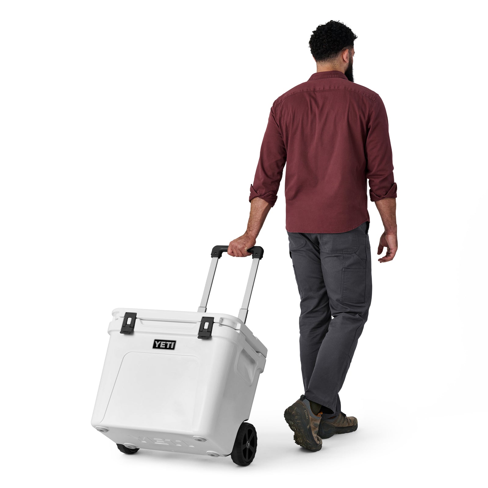 Ein Mann in einem kastanienbraunen Hemd und einer grauen Hose zieht eine weiße YETI® Kühlbox Roadie 60 von YETI vor einem schlichten weißen Hintergrund.