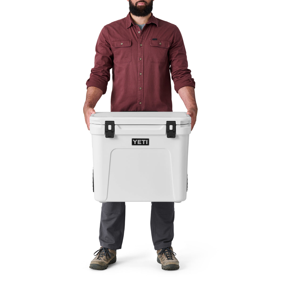 Ein Mann in einem kastanienbraunen Hemd und einer grauen Hose hält eine große weiße YETI® Kühlbox Roadie 60 von YETI vor sich.