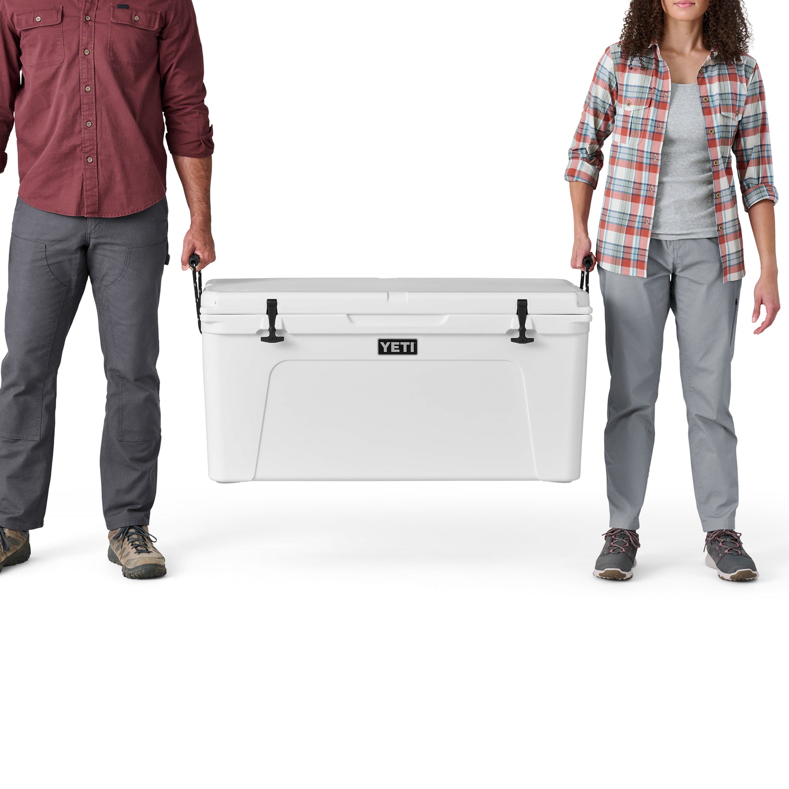 Ein Mann und eine Frau halten die Griffe einer YETI® Kühlbox Tundra 125 - White zwischen sich, wobei nur ihre unteren Hälften sichtbar sind. Der weiße Hintergrund hebt die starke Isolierung der Kühlbox hervor.