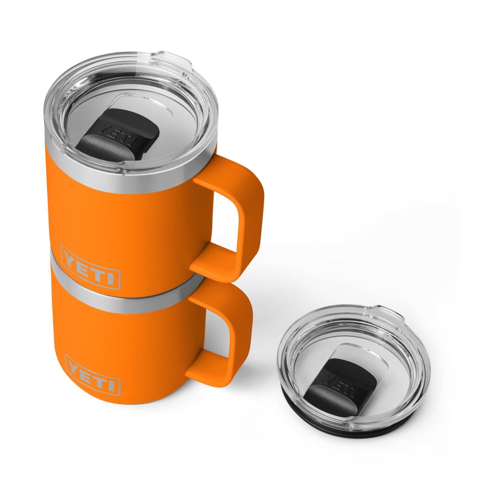 Zwei YETI® Tasse Rambler 10 Oz - King Crab Becher sind mit aufeinander abgestimmten Griffen gestapelt. Die durchsichtigen Deckel, jeweils mit schwarzem Schiebedeckel, ruhen - einer auf einem Becher und der andere daneben auf der weißen Oberfläche.