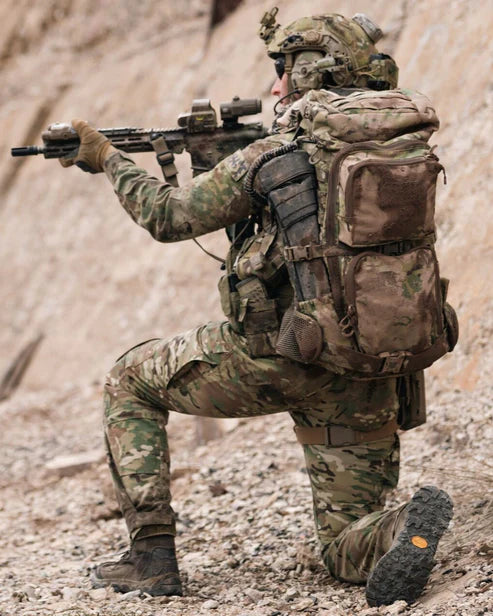 Ein Soldat in Tarnkleidung und mit Helm kniet auf felsigem Boden und richtet sein Gewehr aus. Ausgerüstet für taktische Einsätze, trägt er einen EBERLESTOCK FAC Track-Military Green Rucksack zusammen mit anderer Ausrüstung, die Augen auf ein Ziel vor ihm gerichtet.