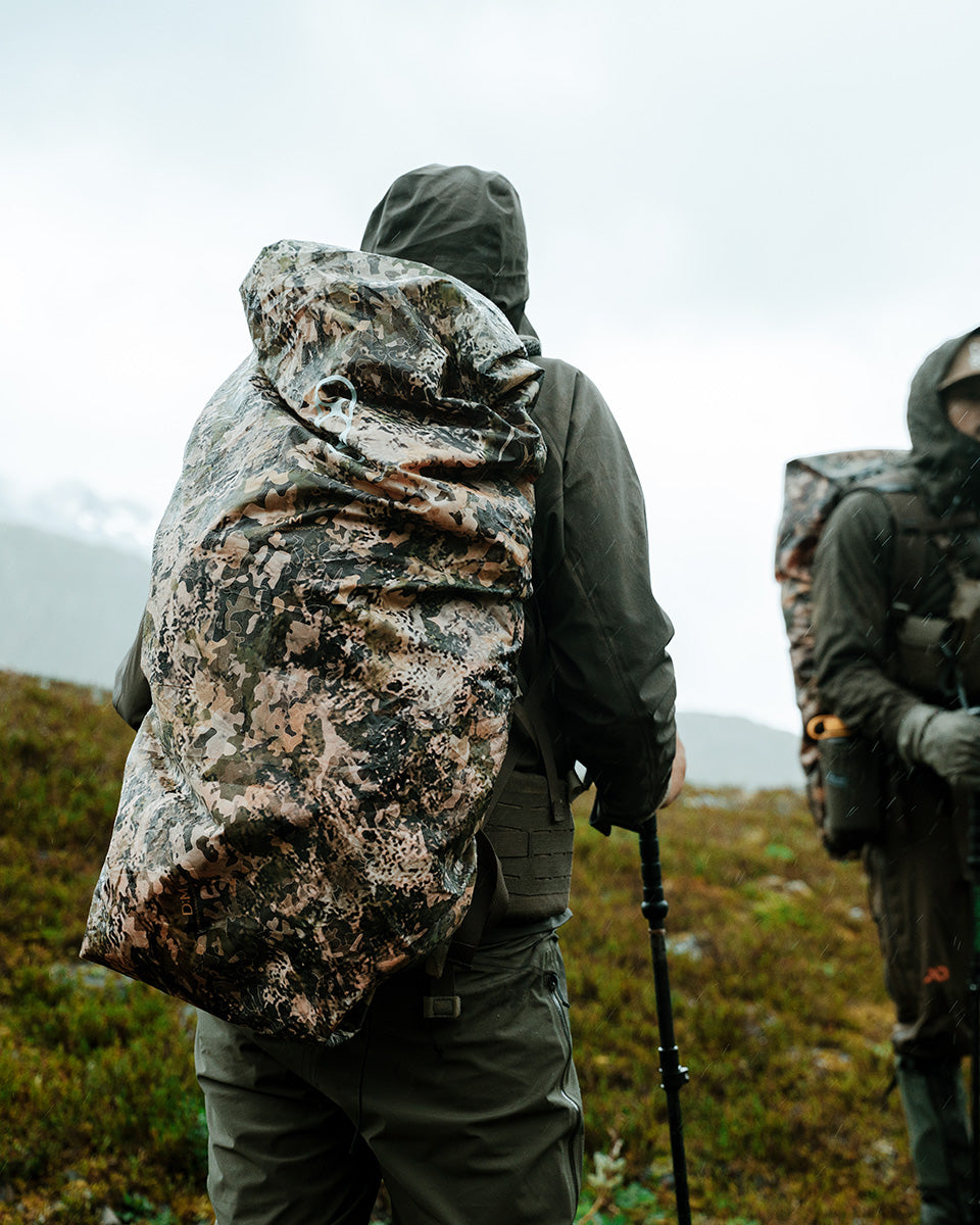 Zwei Wanderer in Tarnjacken mit großen EBERLESTOCK Rucksack-Regenhülle Large wendbar - Mountain / Blaze Orange wandern bei bewölktem und nebligem Wetter mit Trekkingstöcken über einen nebligen, grasbewachsenen Hang.