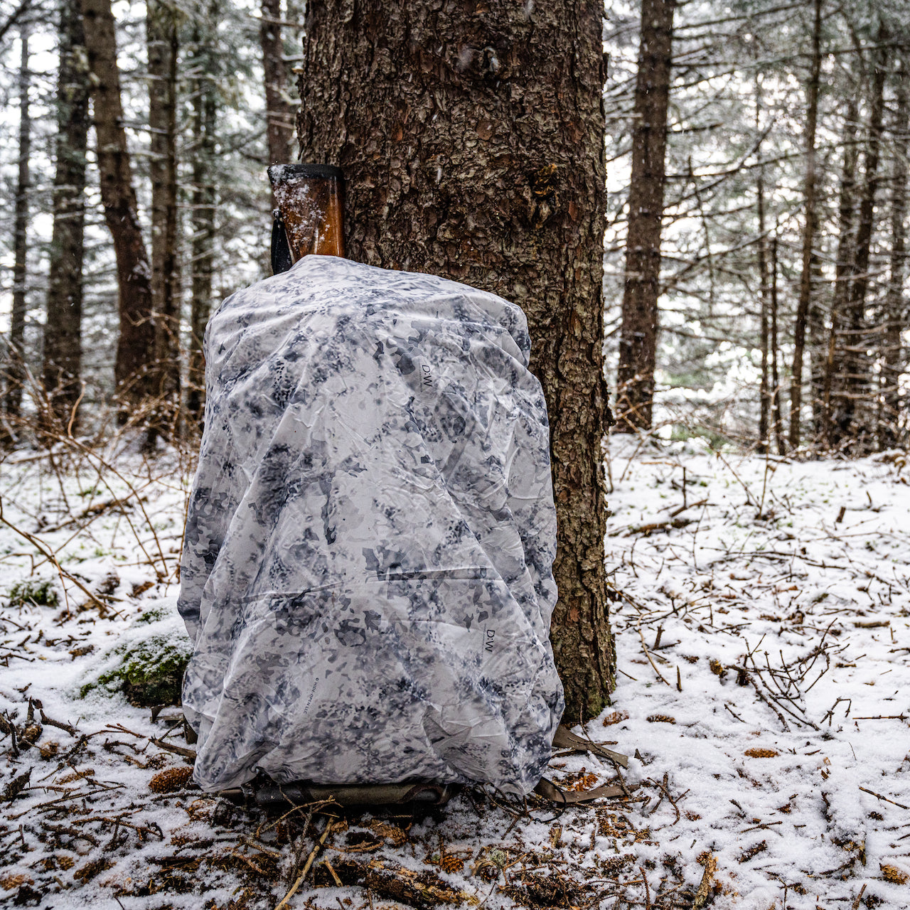 Ein EBERLESTOCK Rucksack mit der Eberlestock Rucksack-Regenhülle Small wendbar - Coyote / D. Winter, Reversible, lehnt an einem verschneiten Baum und fügt sich mit seinem weiß-grau gemusterten Bezug in den Winterwald ein.