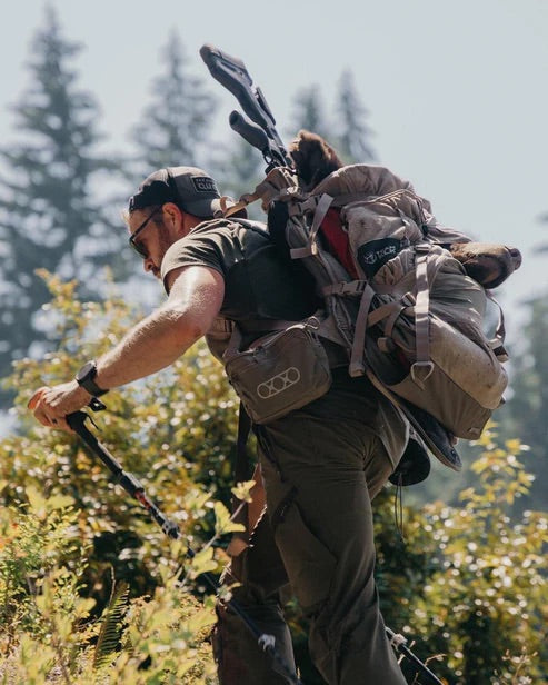 Ein Mann in Outdoor-Ausrüstung und mit Mütze wandert mit einem EBERLESTOCK Team Elk Jagdrucksack - Mountain und einer YETI-Kühlbox auf Trekkingstöcken bergauf durch die grüne Landschaft. Im Hintergrund sind hohe Kiefern und ein heller Himmel zu sehen.
