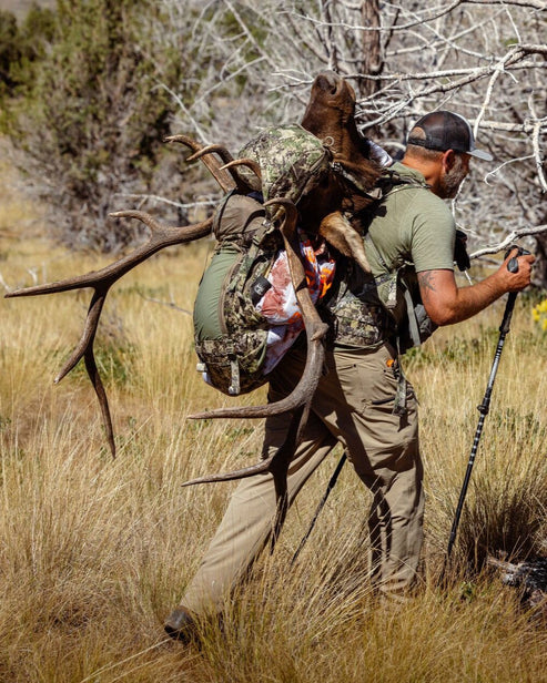 Ein Mann in Tarnkleidung wandert mit dem EBERLESTOCK Team Elk Jagdrucksack - Mountain durch hohes Gras und trägt ein Elchgeweih und eine Wildtasche. Er benutzt einen Trekkingstock bei der Verfolgung von Großwild; im Hintergrund sind Bäume und trockene Äste zu sehen.