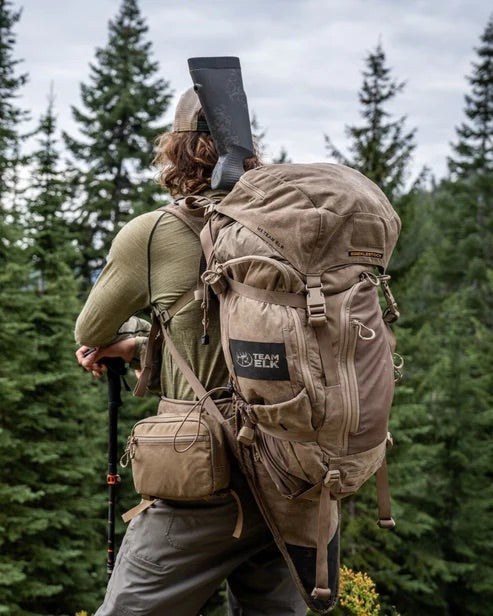 Eine Person in Outdoor-Ausrüstung steht inmitten grüner Kiefern, den Blick abgewandt, mit einem Gewehr auf dem EBERLESTOCK Team Elk Jagdrucksack - Mountain, bereit für die Großwildjagd.
