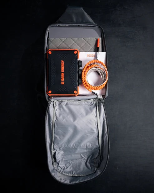 Ein EBERLESTOCK Eberlestock Fade Sling XL, geöffnet auf einer dunklen Oberfläche, enthält ein schwarz-oranges SOLAR EMERGENCY Gerät, eine graue gepolsterte Matte, ein orange-weißes geflochtenes Kabel und eine Broschüre.