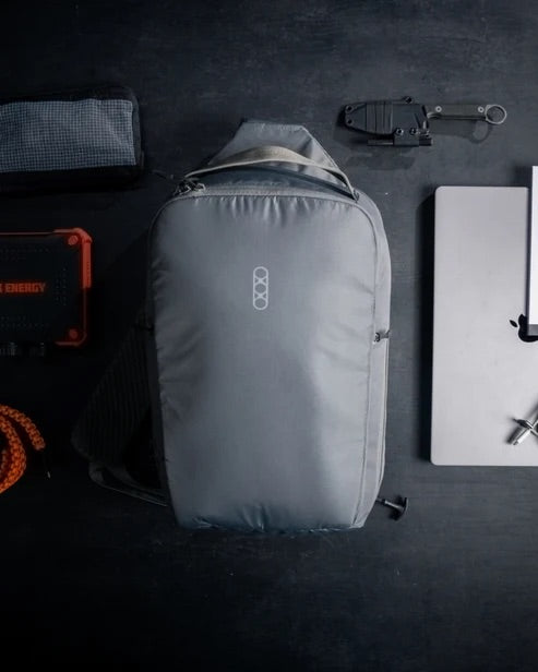 Ein grauer EBERLESTOCK Fade Sling XL Rucksack liegt auf einer dunklen Fläche, umgeben von einem Laptop, einem Multitool, einem orangefarbenen Seil, einer schwarzen Tasche und einer Reißverschlusstasche, die um die taktische Sling-Tasche herum angeordnet sind.