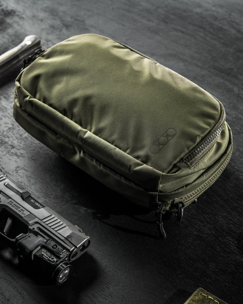 Die olivgrüne EBERLESTOCK Fade Flex Micro Tasche mit Reißverschlussfächern liegt auf einer dunklen Fläche neben einer schwarzen Pistole mit montiertem Licht - ideal für Ihr tägliches Trage-Setup.