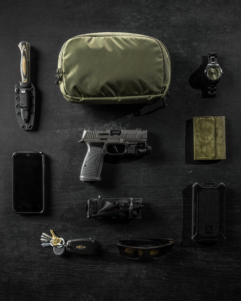 Flache Darstellung einer EDC-Ausrüstung auf einer schwarzen Oberfläche mit einem Messer, einer olivgrünen Umhängetasche, einer Armbanduhr, einem Smartphone, einer Handfeuerwaffe, einer Taschenlampe, einer Geldbörse, Schlüsseln, einer Sonnenbrille und dem EBERLESTOCK Fade Flex Micro Etui.
