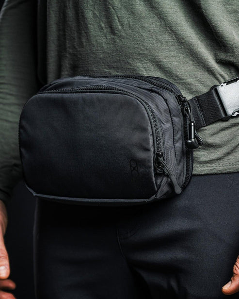 Eine Person in einem dunkelgrünen, langärmeligen Hemd und einer schwarzen Hose trägt eine EBERLESTOCK Fade Flex Micro Tasche mit Reißverschlusstaschen, die mit einer Kunststoffschnalle an der Taille befestigt ist, als Teil ihrer EDC-Ausrüstung.