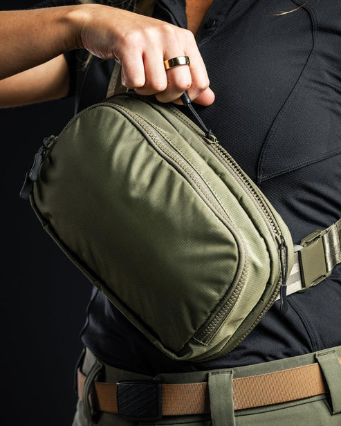 Eine Person mit schwarzem Hemd und hellbraunem Gürtel hält eine olivgrüne EBERLESTOCK Fade Flex Micro in der Hand, greift in den Griff und öffnet ein Fach - ideal für die Organisation Ihrer EDC-Ausrüstung.