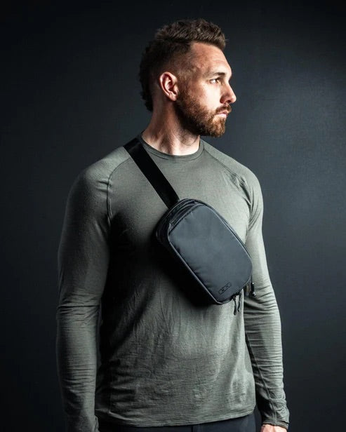 Ein Mann mit kurzen braunen Haaren und einem Bart trägt ein langärmeliges olivgrünes Hemd und eine EBERLESTOCK Fade Flex Micro Tasche. Er steht vor einem dunklen Hintergrund und schaut nach rechts.