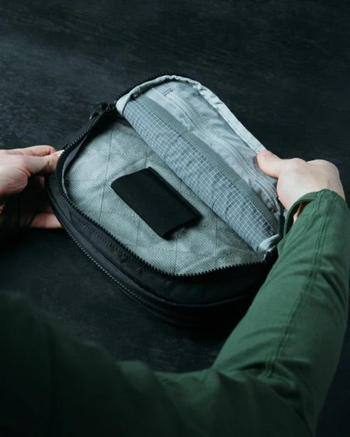 Eine Person mit grünem Ärmel öffnet eine EBERLESTOCK Fade Flex Micro, eine kompakte grau-schwarze Reißverschlusstasche mit einer Innentasche und hellem Futter, die auf einer dunklen Fläche abgebildet ist.