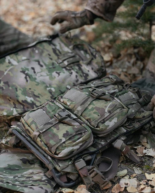 Eine Nahaufnahme einer taktischen Weste von EBERLESTOCK, eines Rucksacks und einer kleinen Eberlestock Rip-Away Medical Pouch mit mehreren Taschen liegt zwischen trockenem Laub, und eine behandschuhte Hand ist in der Nähe der Ausrüstung zu sehen.