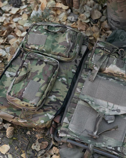 Zwei tarnfarbene taktische Rucksäcke mit mehreren Taschen, darunter eine EBERLESTOCK Rip-Away Medical Pouch small, und Klettverschlüssen werden auf den mit trockenen Blättern und Zweigen bedeckten Boden gelegt.