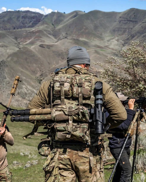 Eine Person in Tarnkleidung, die eine EBERLESTOCK Rip-Away Medical Pouch Large und einen Rucksack trägt, steht im Freien mit einem Gewehr und einem Stativ inmitten von Bergen, dazu gesellen sich andere mit Kameraausrüstung.