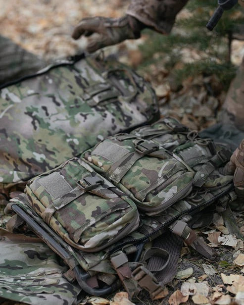 Eine Nahaufnahme der militärischen Tarnkleidung zeigt eine taktische Weste mit Taschen, eine EBERLESTOCK Rip-Away Medical Pouch Large und einen Rucksack auf trockenem Laub, während eine behandschuhte Hand nach der medizinischen Ausrüstung greift.