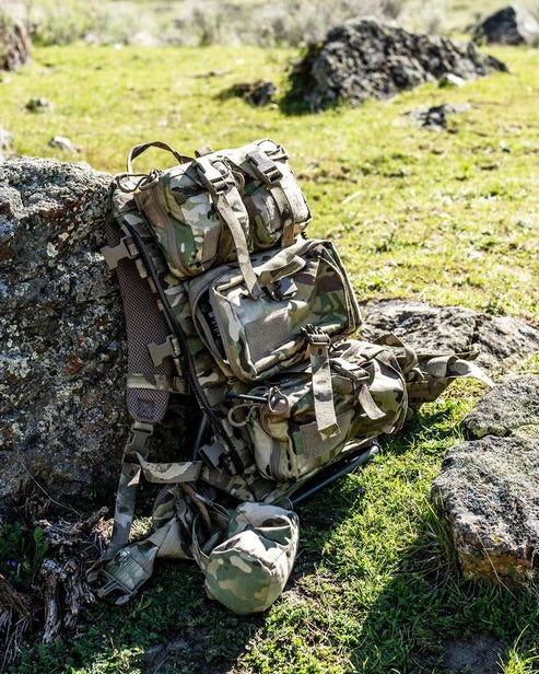 Eine EBERLESTOCK Rip-Away Medical Pouch Large ruht an einem getarnten Rucksack auf grasbewachsenem Terrain im Sonnenlicht, umgeben von Felsen und Grünzeug.