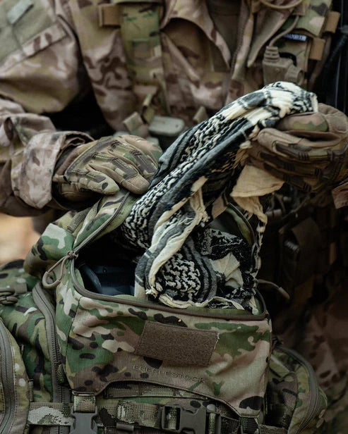In Tarnkleidung und mit Handschuhen packt jemand den taktischen Rucksack EBERLESTOCK Mission Lid mit zusätzlichem Stauraum ein oder aus und hält dabei einen schwarz-weiß gemusterten Schal in der Hand.
