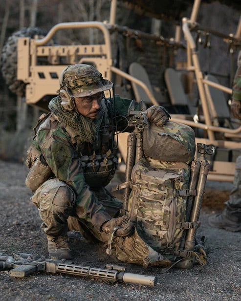 Ein Soldat in Camouflage kniet und verstellt einen EBERLESTOCK Mission Lid für zusätzlichen Stauraum an seinem Rucksack. Dahinter tauchen Militärfahrzeuge und ein weiterer Soldat auf, die das modulare Eberlestock Mission System bei der Feldausbildung hervorheben.