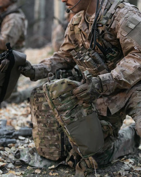 Ein Soldat in Camouflage kniet im Wald mit einem EBERLESTOCK Rucksack, der mit Eberlestock Mission Wings für zusätzlichen Stauraum ausgestattet ist. Er trägt Schutzhandschuhe, und an seiner Weste sind eine Kommunikationsausrüstung und seitliche Erweiterungsmodule befestigt.
