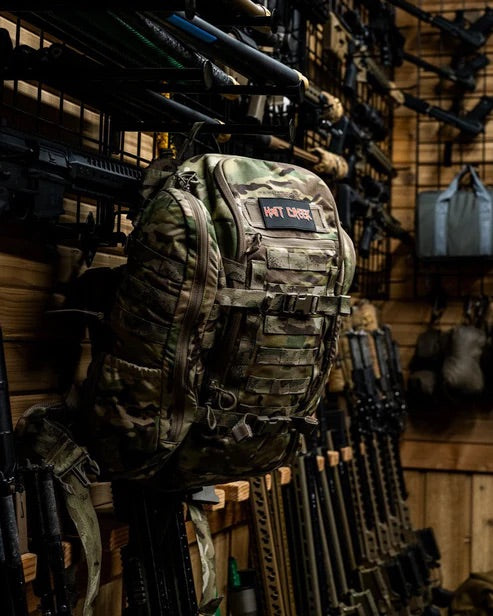 Ein EBERLESTOCK Mission Wings Militärrucksack in Tarnfarbe mit zusätzlichem Stauraum und seitlichen Erweiterungsmodulen hängt in einem Lagerraum mit Holzwänden an der Wand, umgeben von Gewehren und taktischer Ausrüstung.
