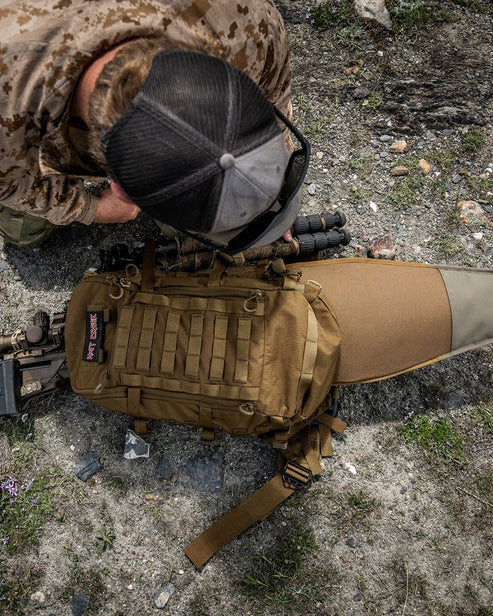 Eine Person in Tarnhemd und schwarzer Netzmütze kniet auf felsigem Boden neben einem EBERLESTOCK Eberlestock Mission Assault Pack und einem Gewehr in einem Koffer, von oben gesehen.
