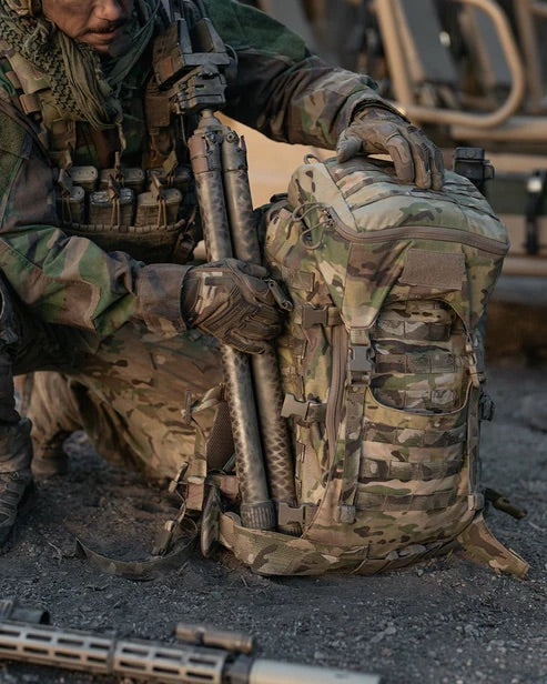 Ein Soldat in Tarnkleidung und taktischer Weste kniet vor einem EBERLESTOCK Mission Assault Pack mit modularem Design, MOLLE-Gurtband und befestigter Ausrüstung.
