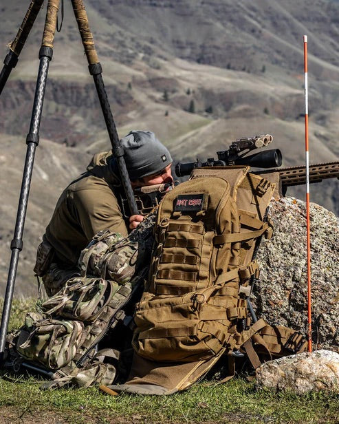 Eine Person in Outdoor-Ausrüstung liegt auf dem Bauch und zielt mit einem Gewehr auf einem Stativ neben einem EBERLESTOCK Mission Assault Pack und Tarnkleidung mit einer gestreiften Stange in der Nähe eines großen Felsens in den Bergen.