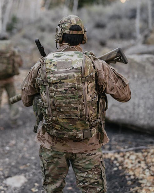 Eine Person in Tarnkleidung und einem EBERLESTOCK Mission Assault Pack steht im Freien und hält ein Gewehr, das von der Kamera weg zeigt. Im felsigen, bewaldeten Hintergrund ist links eine weitere verschwommene Figur zu sehen.