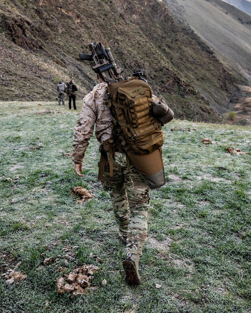 Ein Soldat in Tarnkleidung überquert einen grasbewachsenen, frostigen Hügel mit einem EBERLESTOCK Mission Assault Pack und einem umgeschnallten Gewehr. Zwei Kameraden gehen in der Ferne voraus.